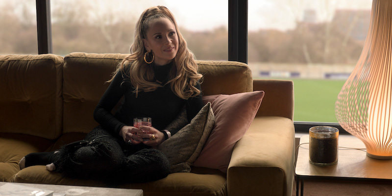 Keeley played by Juno Temple sits on a leather couch with a drink